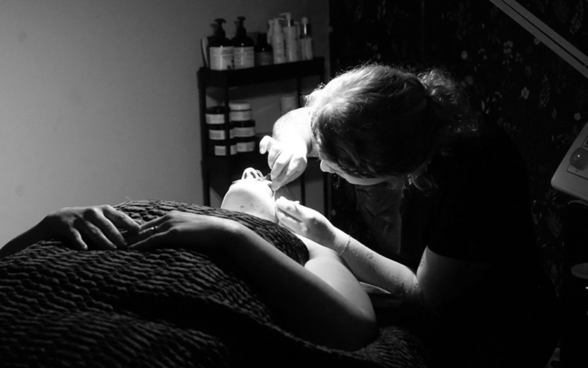Person receiving facial treatment; esthetician's hands, close-up, working on face. Black and white, spa-like setting.