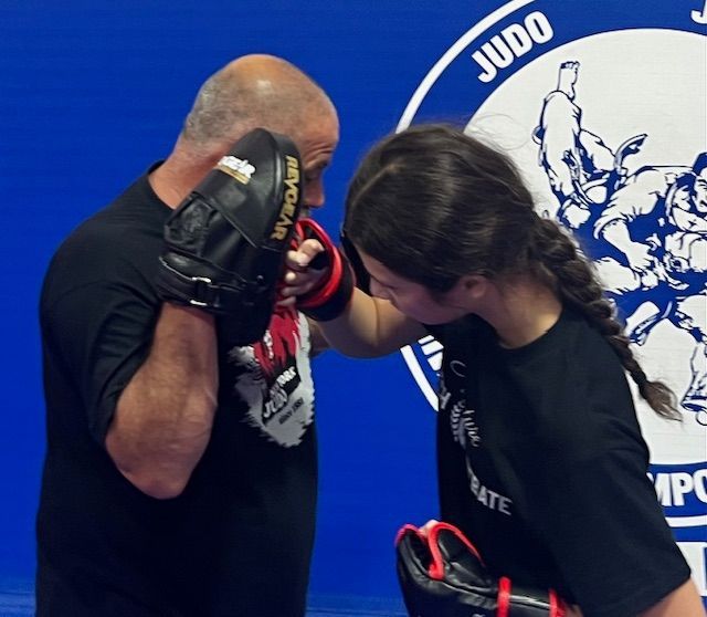 A coach holds a black punch mitt for a person wearing red-and-black gloves during a martial arts training session.