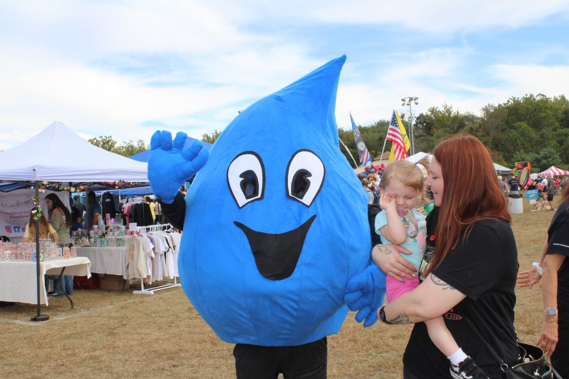 A blue, smiling water drop mascot waves while standing next to a person holding a small child at an outdoor event.