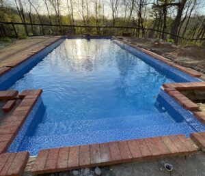 A rectangular backyard pool with blue tiled steps and a red brick border, surrounded by trees at sunset.