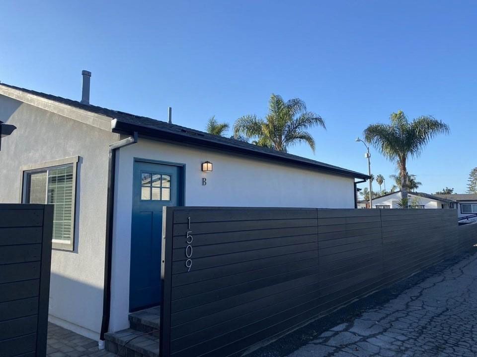Small white house with blue trim and black fence under a clear sky, with palm trees in the background