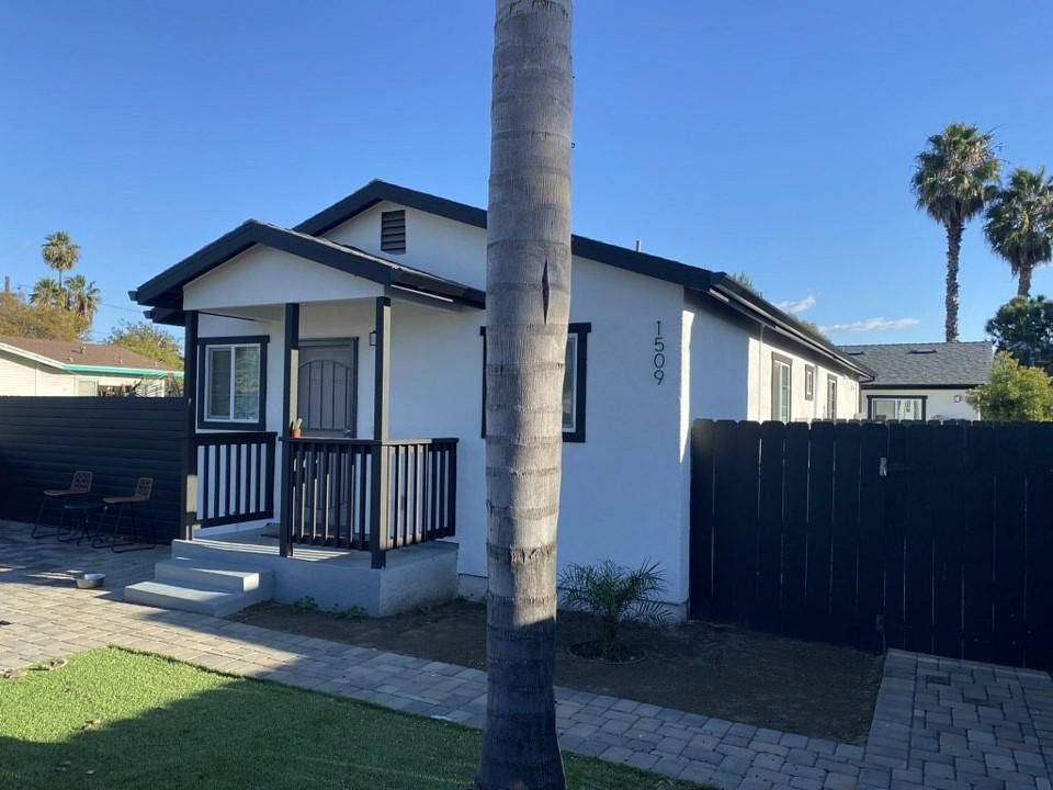 Small white house with a black porch, palm tree in front, and a fenced yard under a clear blue sky