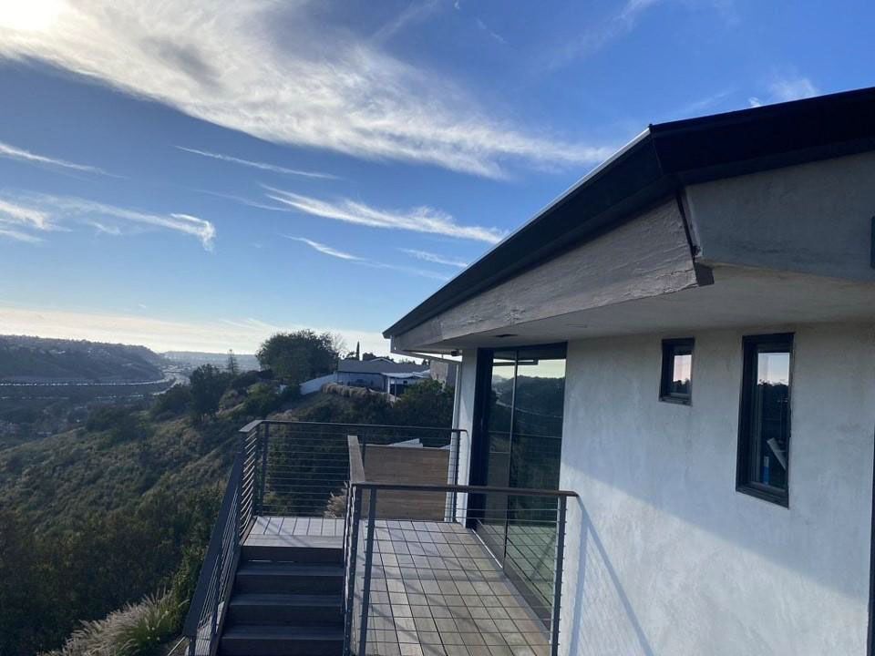 Rooftop terrace with metal railing beside a white house, overlooking a hillside under a blue sky