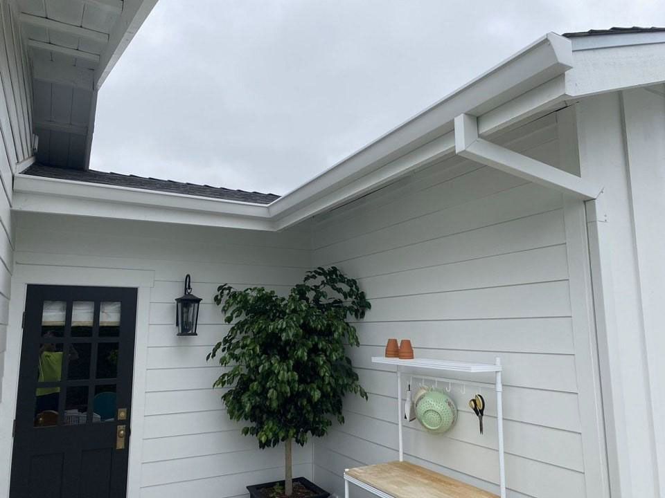 White house exterior with black door, wall lantern, potted tree, and decorative mailbox under an eave