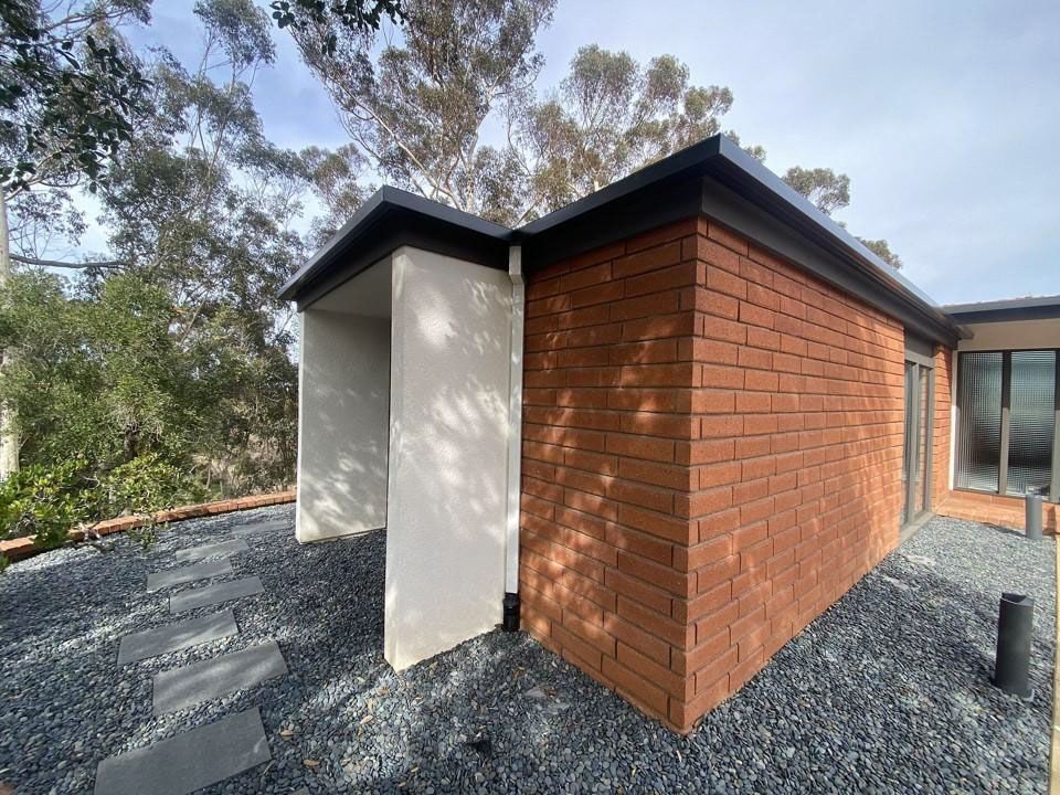 Modern brick house exterior with a white side wall, black roof, and gravel landscaping among trees