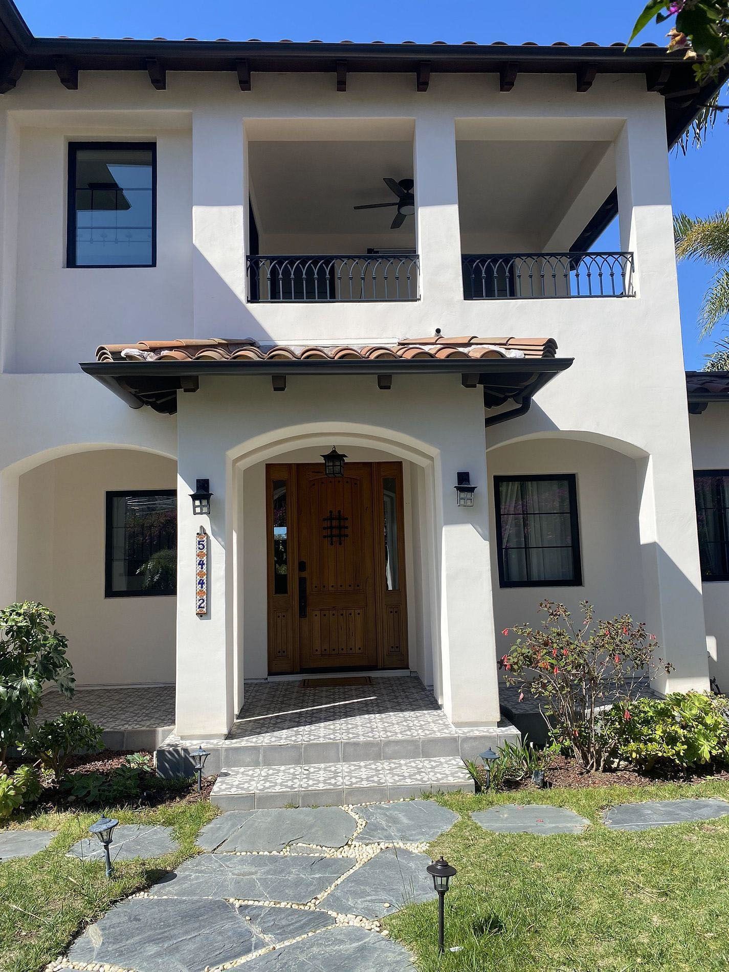 Two-story stucco house with arched entry, wooden front door, balcony, and stone path in front garden