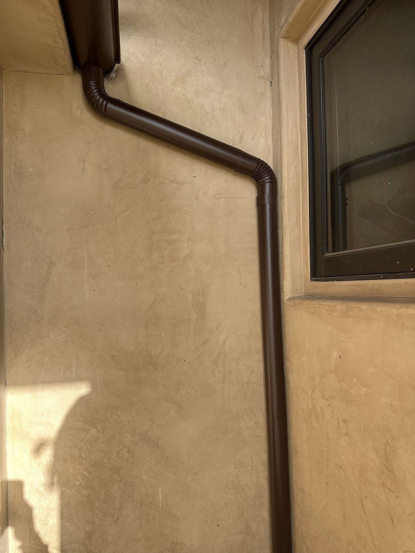 Brown stucco wall with a dark pipe and a window in sunlight