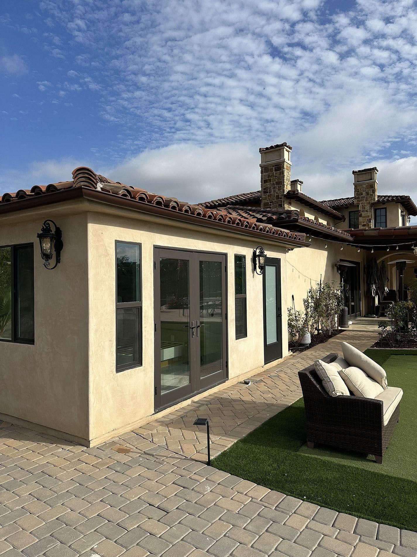 Patio beside cream stucco house with glass doors, wicker sofa, and blue sky.