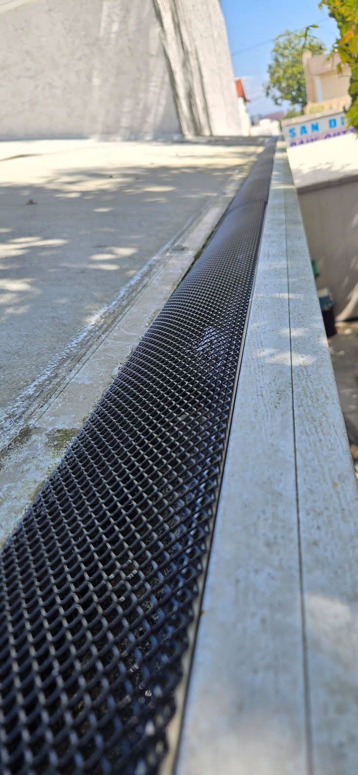 Metal drainage grate along a concrete curb beside a sloped roadway in bright sunlight