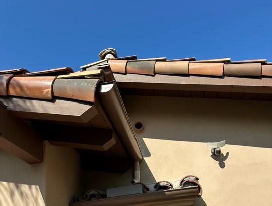 House roof with brown tiles, downspout, and wall-mounted security camera under a clear blue sky