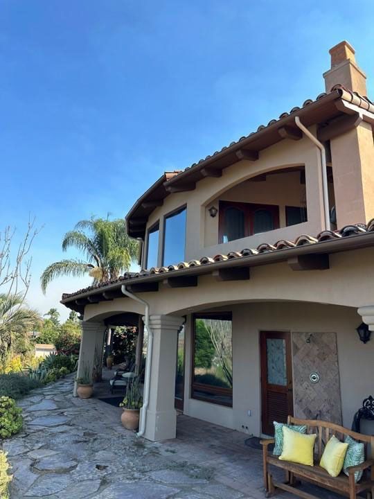 Spanish-style stucco house with stone path, tiled roof, palm tree, and covered porch with outdoor chairs
