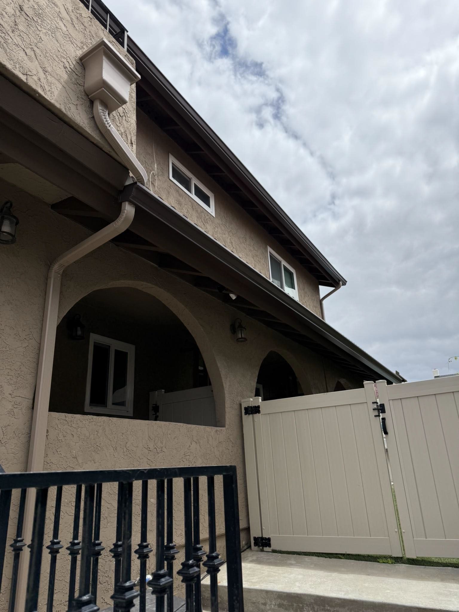 Courtyard view of a beige house with arched porch, white fence, and cloudy sky