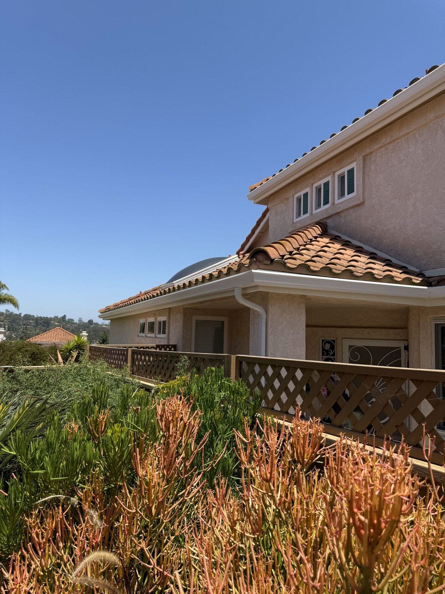 Suburban house with beige stucco, terracotta roof, and a wooden fence behind tall dry grass under a blue sky