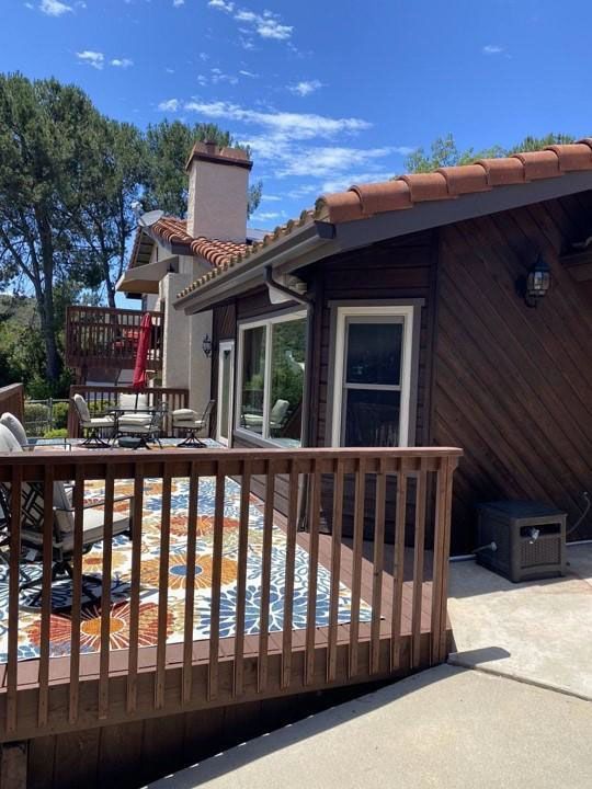 Wooden deck with patterned rug, sliding glass doors, and a terracotta-roofed house under a blue sky