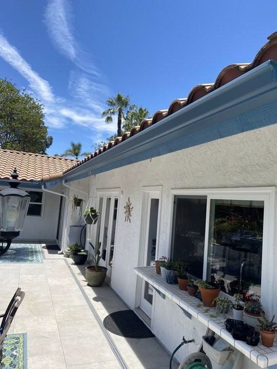 Sunlit white house patio with blue trim, potted plants, windows, and a tiled roof under a clear sky