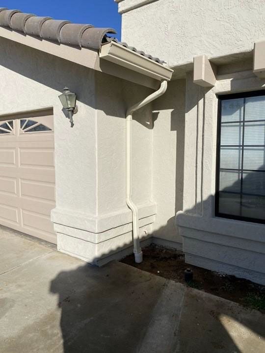 Stucco house corner with roof gutter, garage door, window, and concrete driveway in sunlight