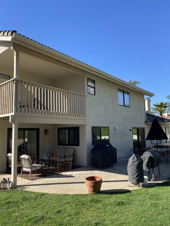 Two-story beige house with balcony, patio furniture, grill, and lawn under a clear blue sky