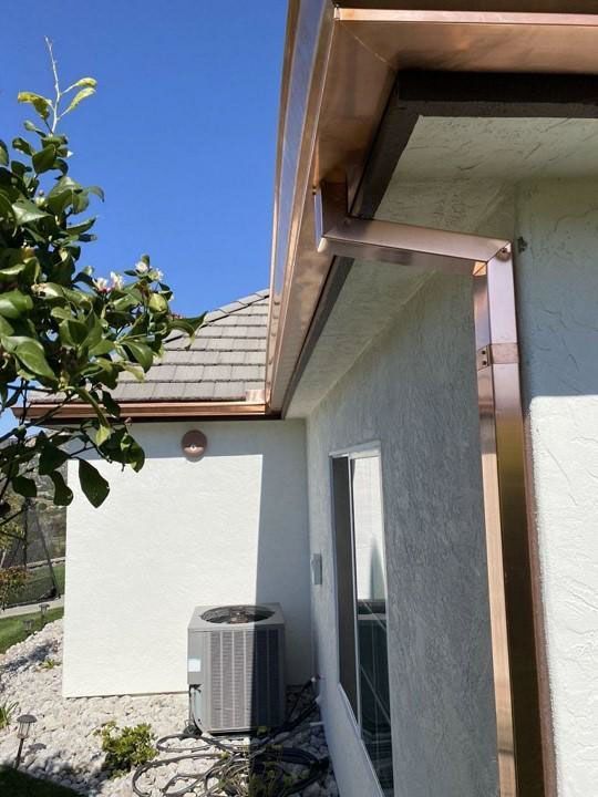 Side of a house with white stucco walls, roof eaves, an air conditioner, and a tree under a blue sky