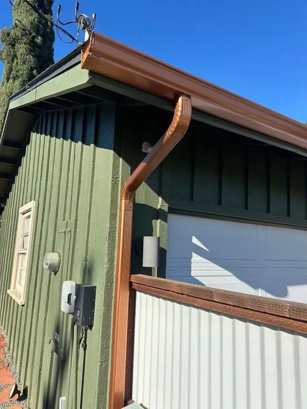 Green house corner with brown downspout, white garage door, and corrugated metal fence in sunlight.