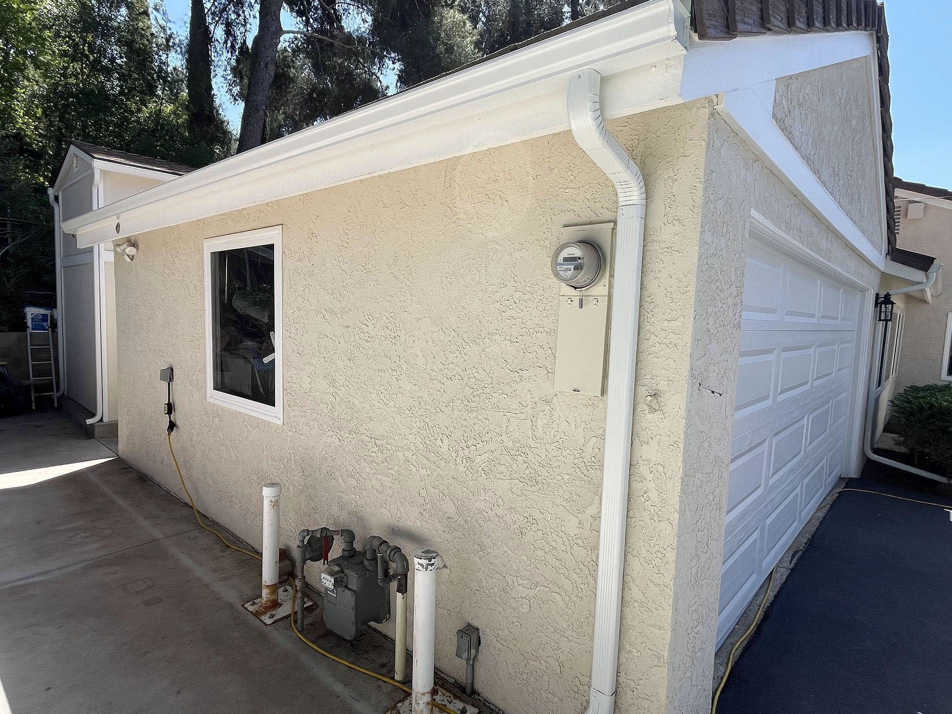 Side view of a beige stucco house with a white garage door, window, and utility meters near the wall