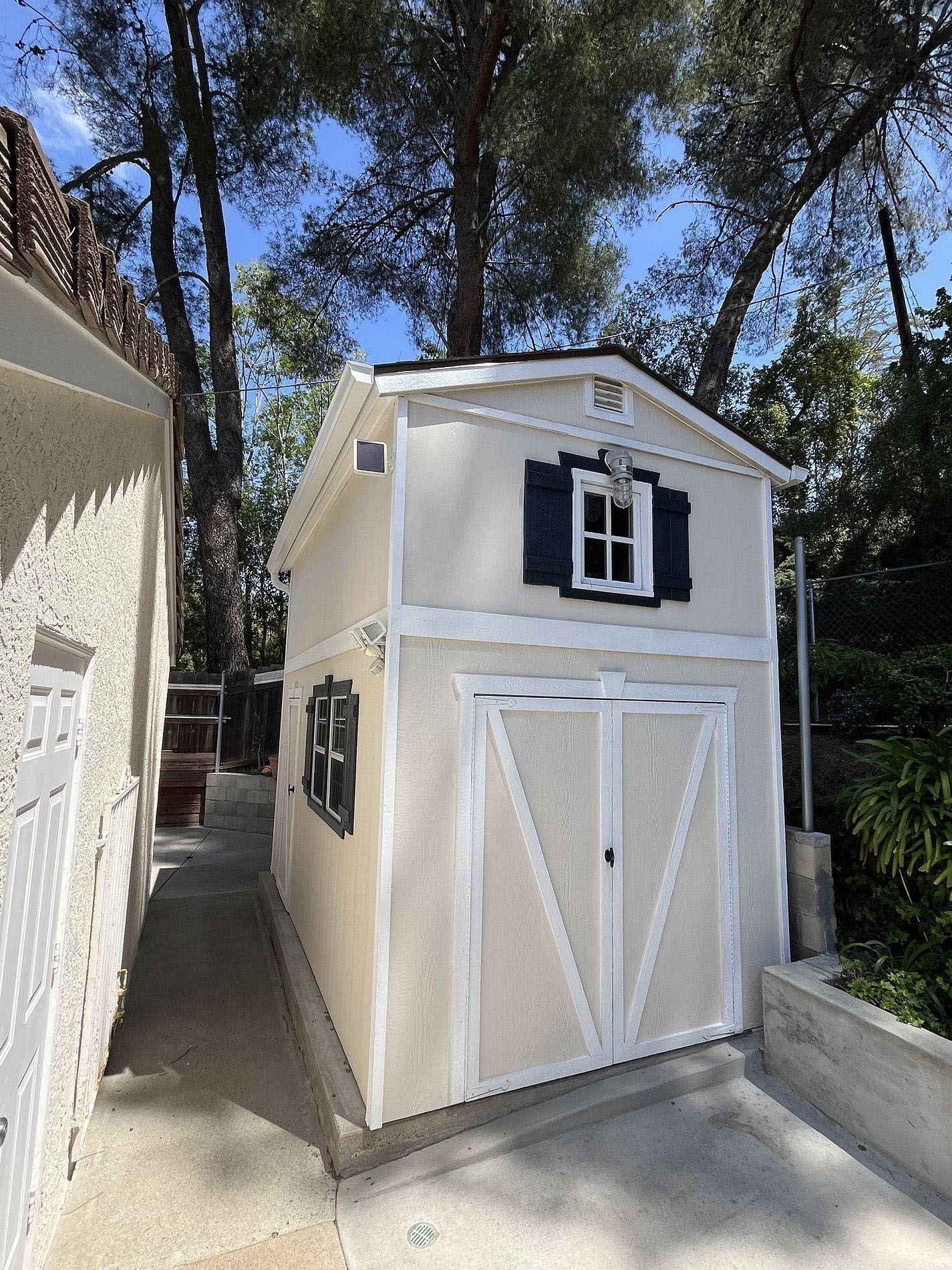 Small beige playhouse with white trim and black shutters beside a house in a wooded yard