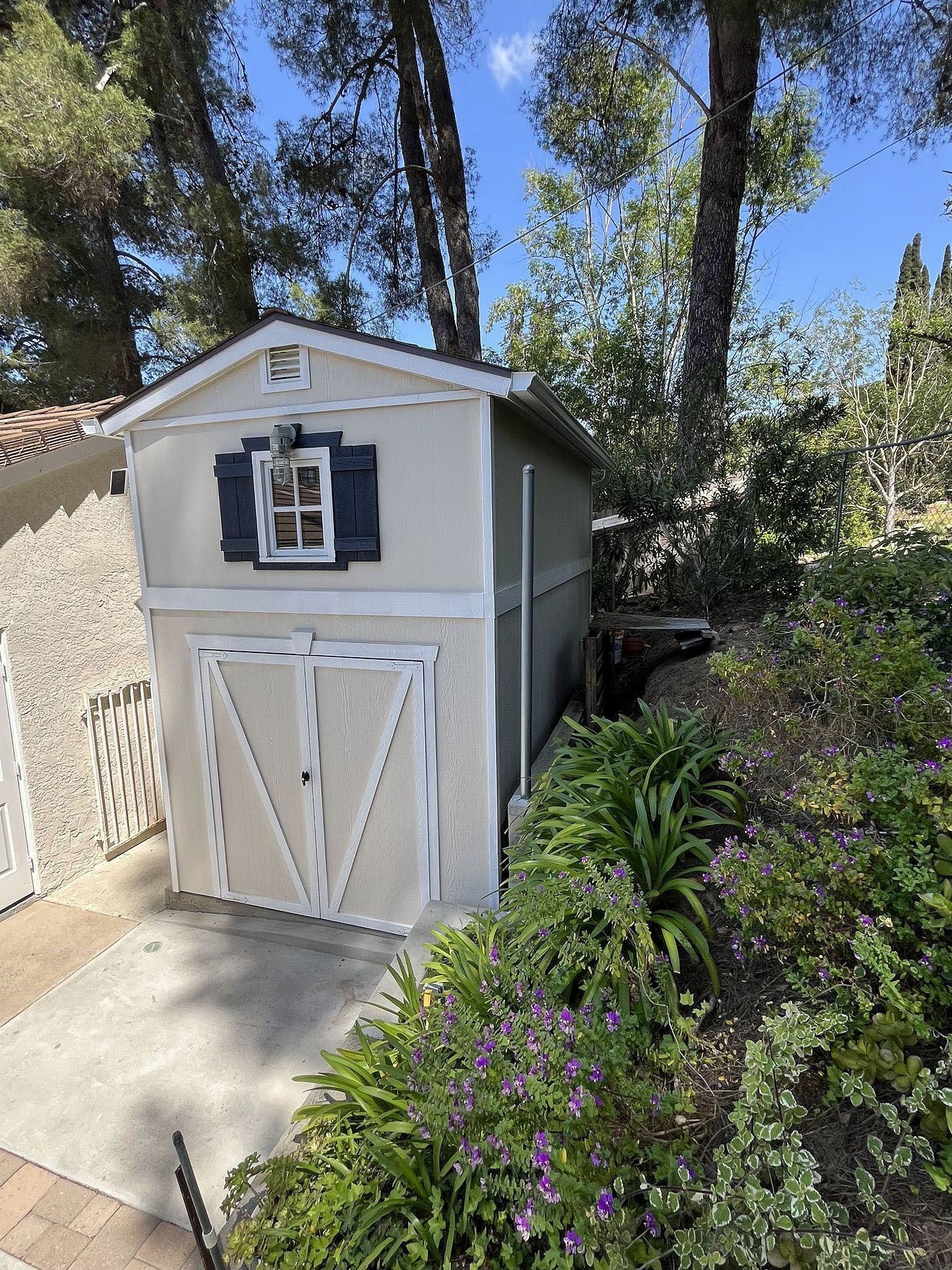 Small gray garden shed with white trim beside a concrete path and flowering plants under tall trees.