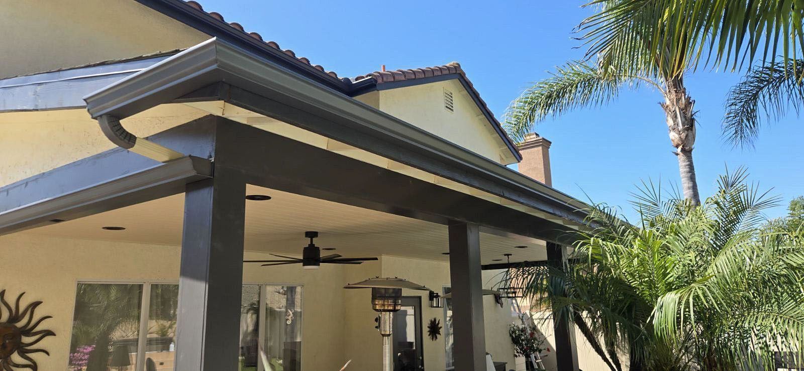 Covered patio with ceiling fan and tiled roof beside a house, framed by palm trees and blue sky