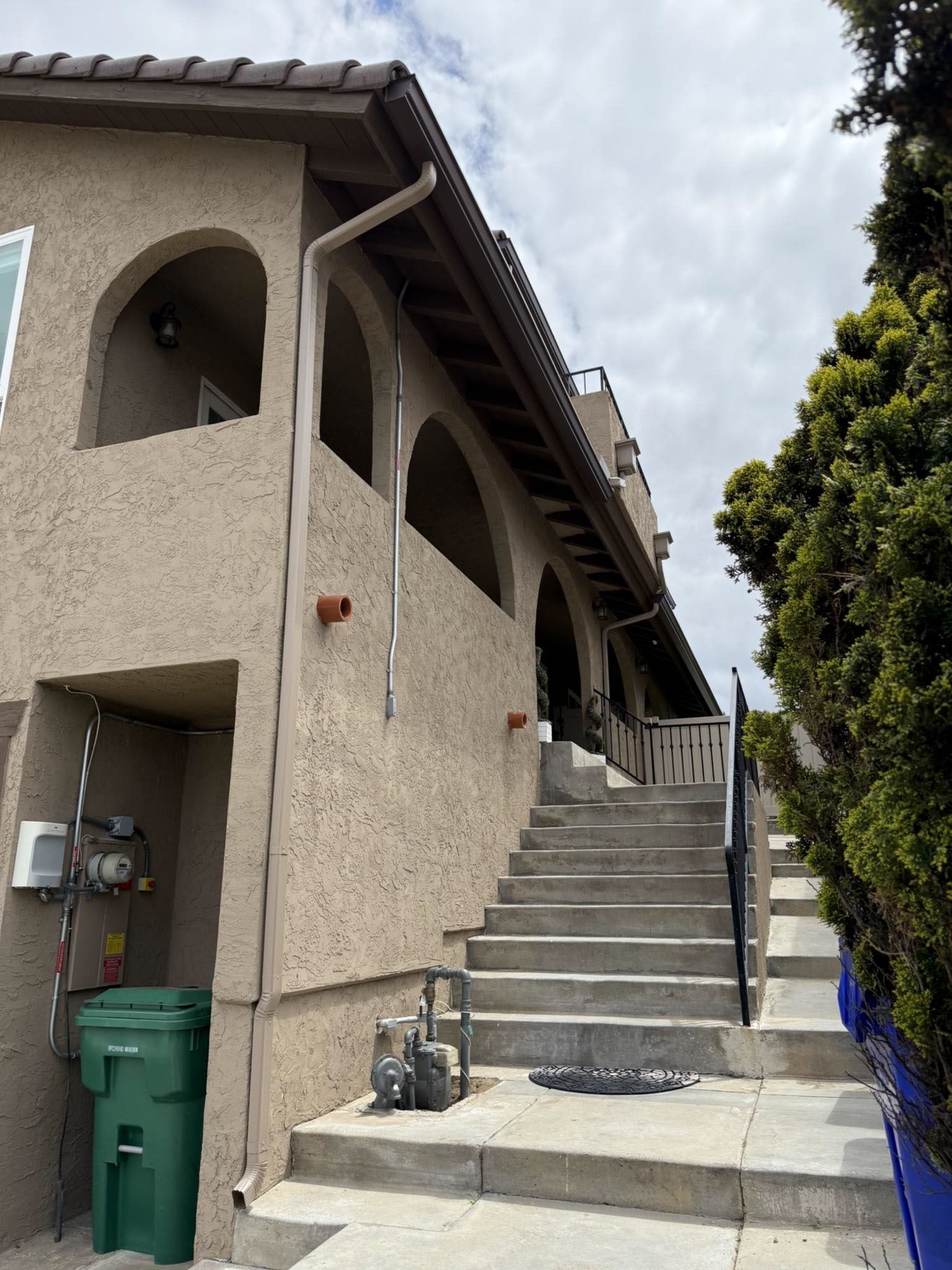 Exterior staircase beside a beige stucco building, with green trash bins and a tree on the right.