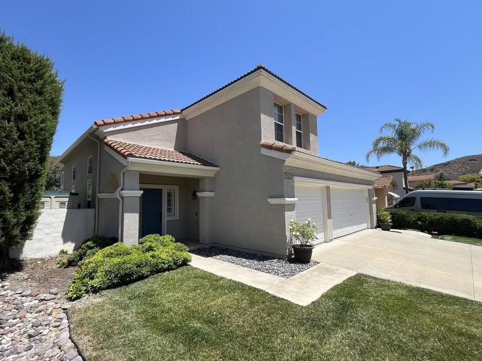 Suburban two-story house with a beige exterior, tiled roof, driveway, and trimmed front lawn under a clear blue sky