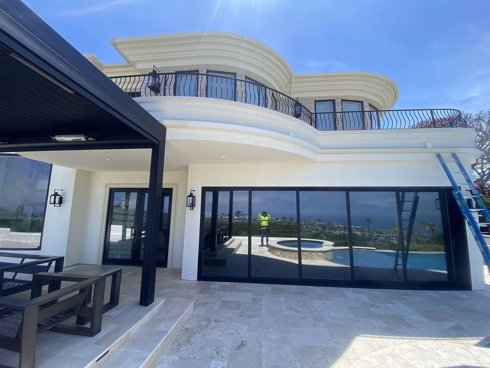 Modern white villa with curved balcony, glass doors, patio seating, and sea view under a clear blue sky