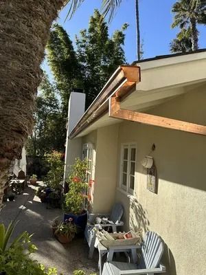 Sunny patio beside a cream house with white chairs, potted plants, and a wooden awning.