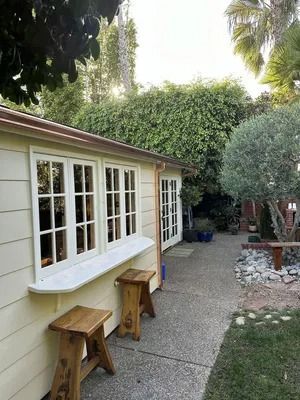 Small yellow house exterior with white windows, wooden stools, and a gravel path beside greenery.