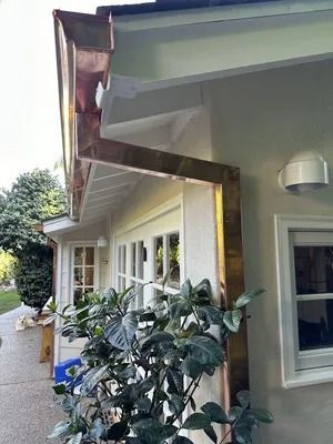 Damaged roof gutter above a house exterior with white trim and a window, next to shrubs and a driveway