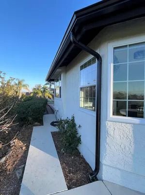 Exterior walkway beside a white stucco house with windows, black roof trim, and shrubs under a blue sky