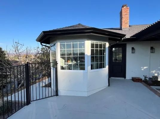White exterior house with a bay window and black railing on a sunny patio with snow nearby