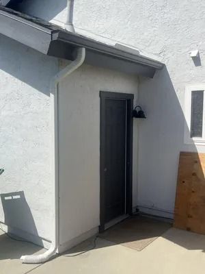 White exterior wall with a dark door, small wall light, and wooden board leaning nearby under a roof eave.