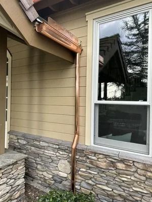 Copper downspout running from roof to stone-and-beige siding beside a large window