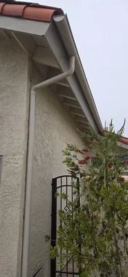 Exterior of a house with gray stucco walls, white roof trim, red tile roof, and a black metal gate beside shrubs