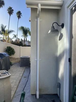 Narrow side yard with a wall-mounted light, white downspout, pavers, grill, and palm trees beyond the fence