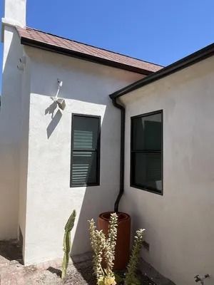 White stucco house corner with two dark windows, red roof trim, and potted plants in bright sunlight