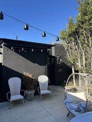 Outdoor patio with black wall, string lights, white chairs, and a wicker hanging chair beside a tree