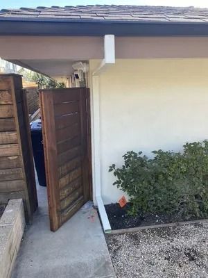 Open wooden gate beside a house wall, with a small garden and gravel path.