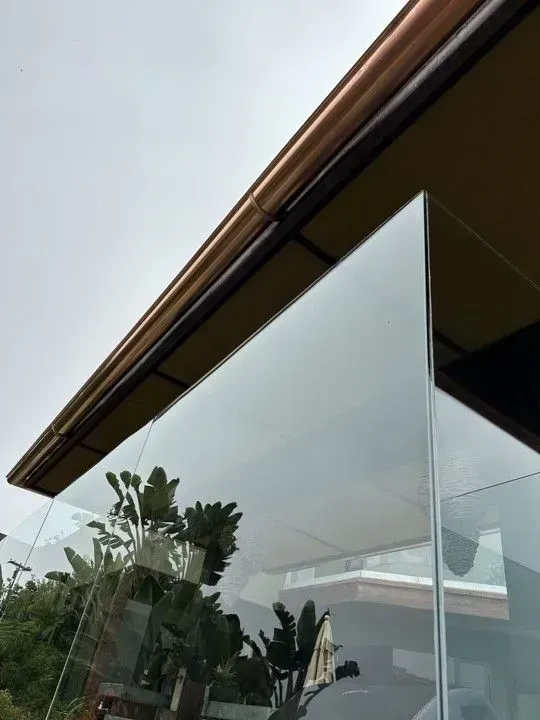 Modern glass patio enclosure with potted plants under a brown roof.