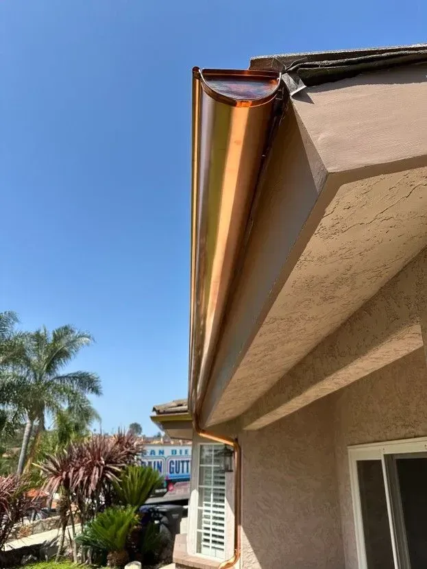 Copper rain gutter along a beige stucco house roofline against a blue sky.