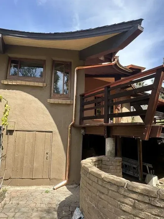 Adobe-style building with curved roof, wooden balcony, and stone wall in bright daylight