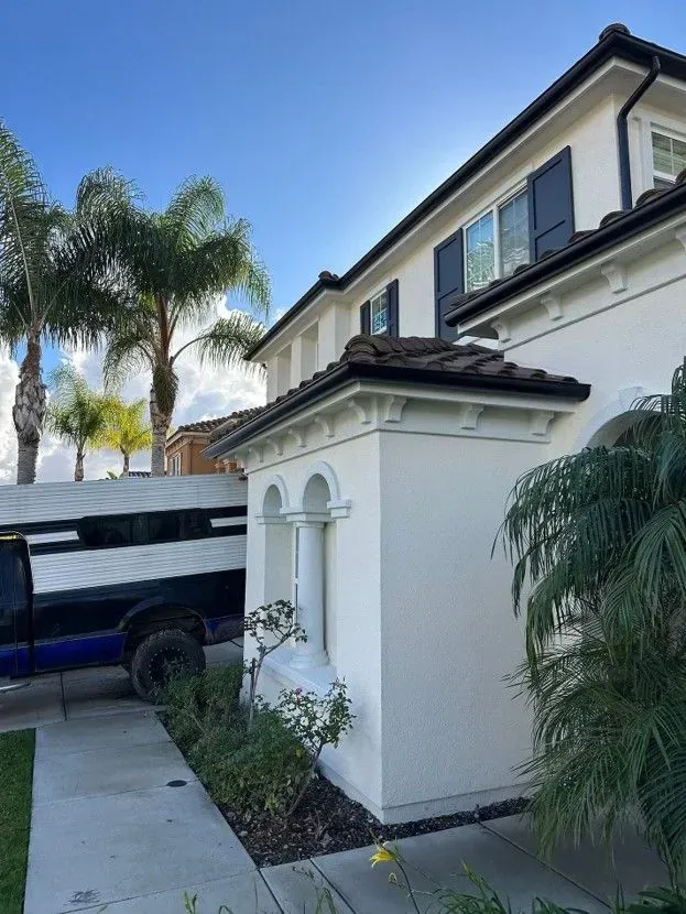 White two-story house with black trim and a small side structure beside a driveway and palm trees.