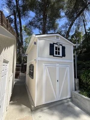 Small cream storage shed with white doors and black shutters beside a house in a wooded driveway.