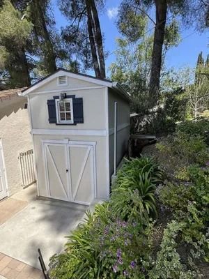 Gray garden shed with white trim and blue shutters beside a driveway and greenery.