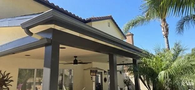 Covered patio with dark trim, ceiling fan, and palm trees beside a stucco house