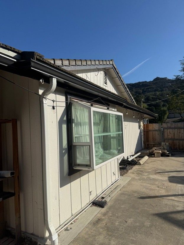 Sunlit side of a white house with green window shutters, concrete patio, and mountain view under a blue sky